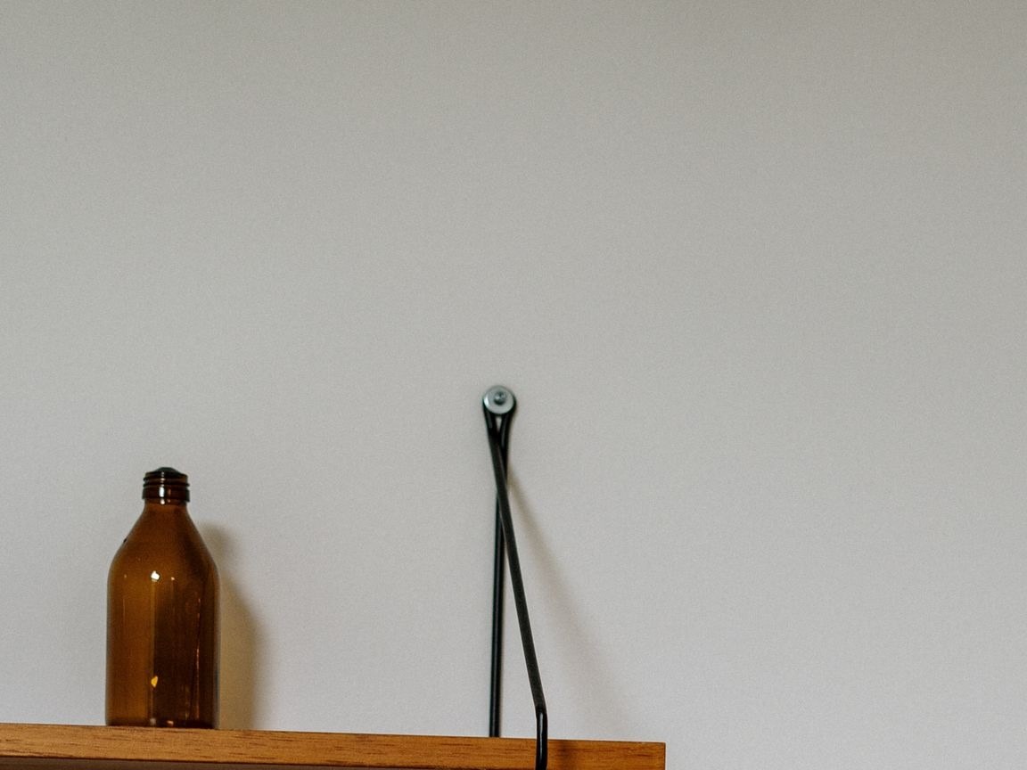 A small amber glass jar sits on the left end of a light wooden wall shelf; the rest of the board is bare.