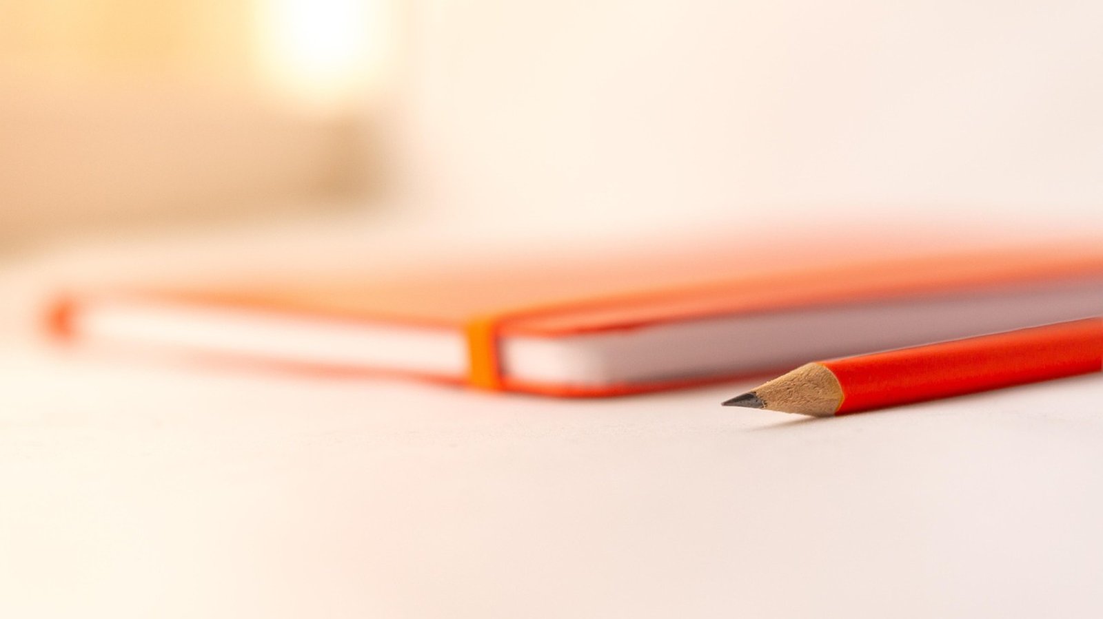 A closed orange notebook lies out of focus on a pale table, with a red pencil in the foreground and its point turned toward the left.