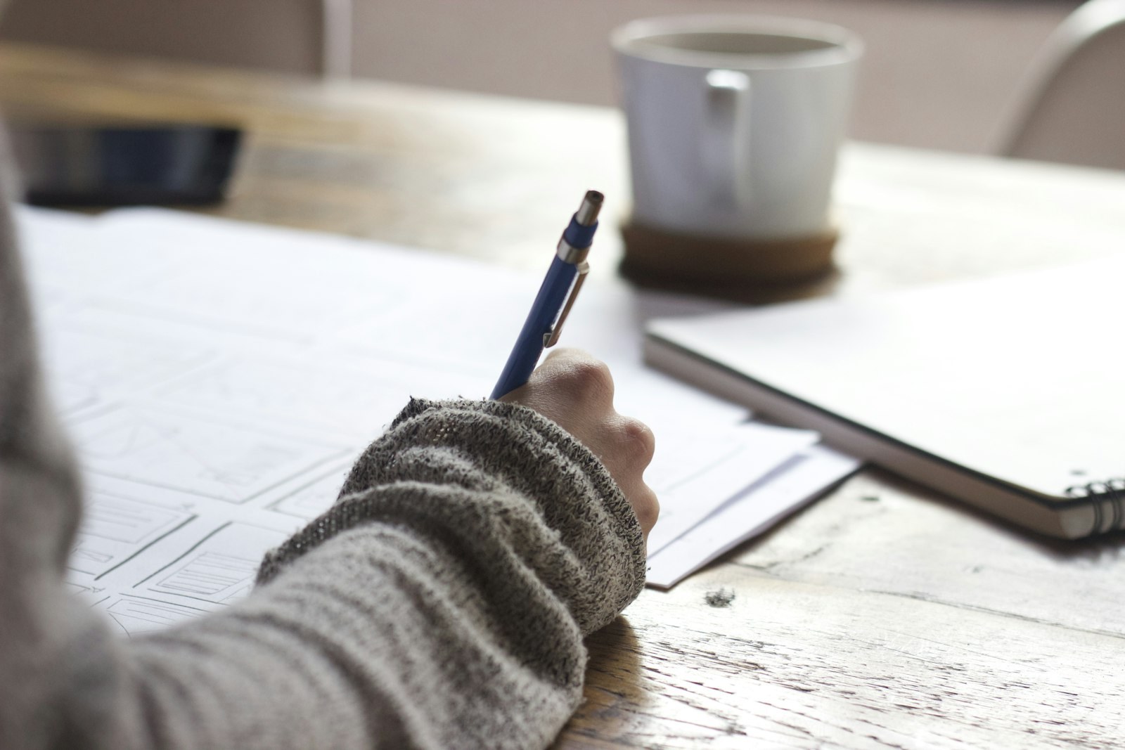A loose white sheet on a wooden table shows faint pencil rectangles, ticks, and illegible scribbles; a hand holds a blue mechanical pencil on the page.
